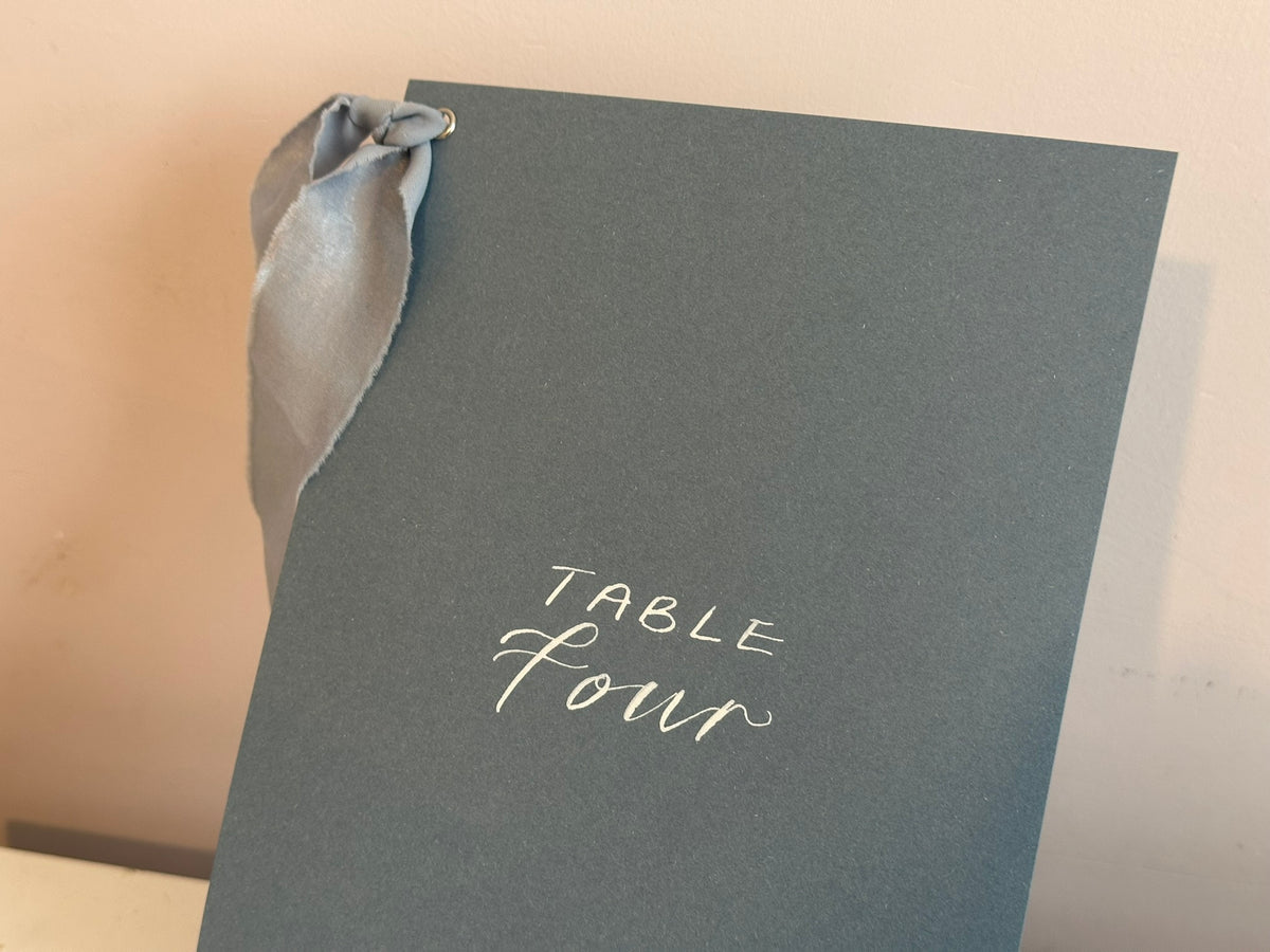 Mixed lettering wedding table number sign on dusky blue card with chiffon ribbon and silver calligraphy