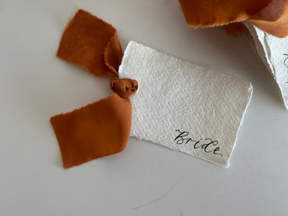 Close-up of an ivory handmade paper wedding place card with ochre chiffon ribbon and gold eyelet