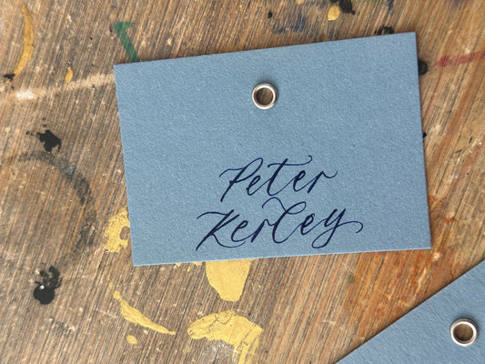 A close-up of a blue paper card with elegant NAVY cursive calligraphy reading 'Peter Jenner' and a small metal grommet on the right side, placed on a rustic wooden surface.