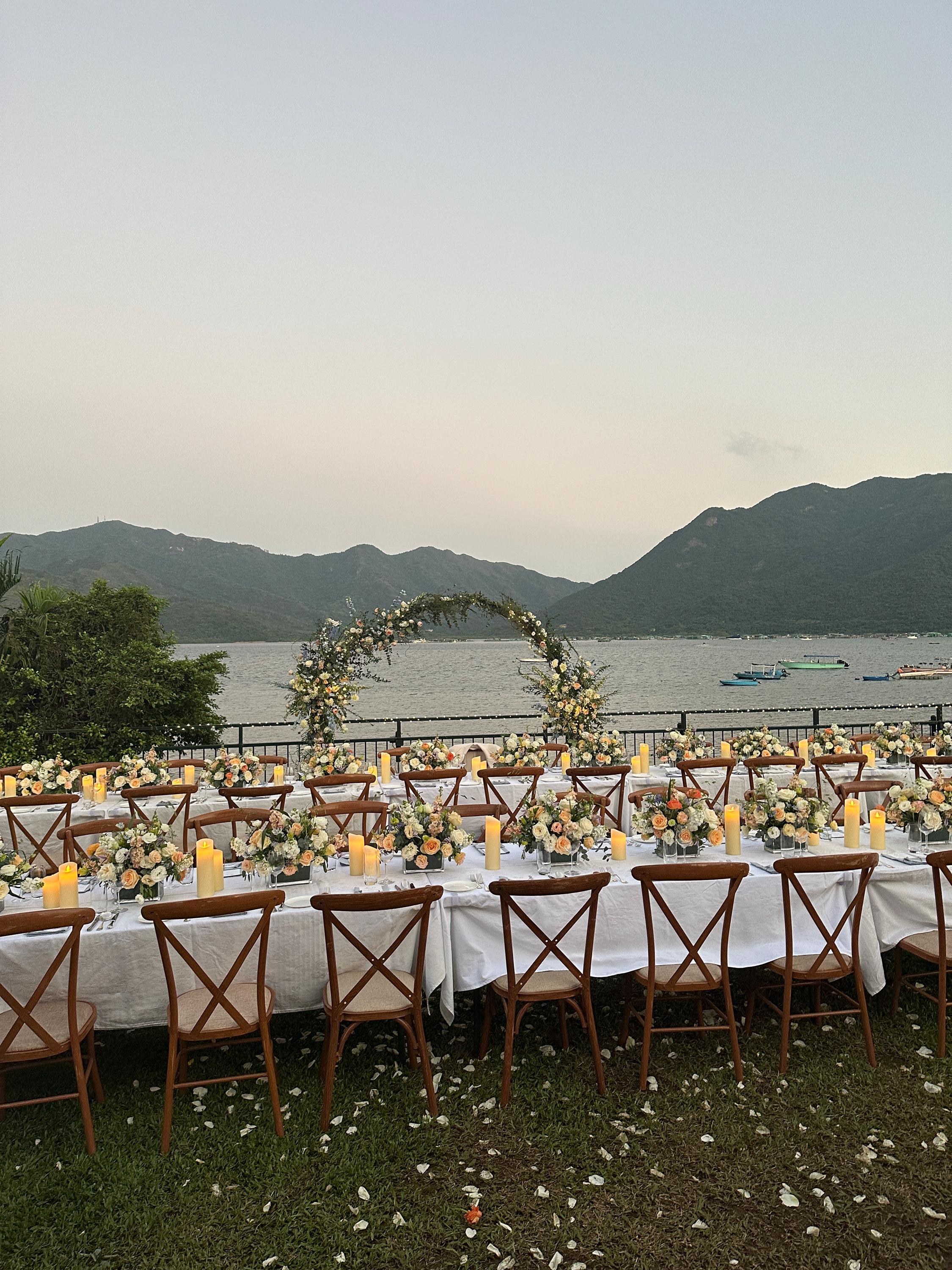 Outdoor wedding setup with tables, chairs, and floral arch by a lake with mountains in the background.