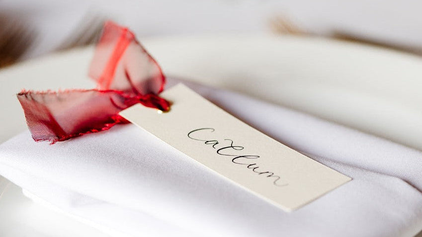 Close up of handwritten calligraphy place card with gold eyelet and organza ribbon, textured paper, soft romantic lighting, shallow depth of field