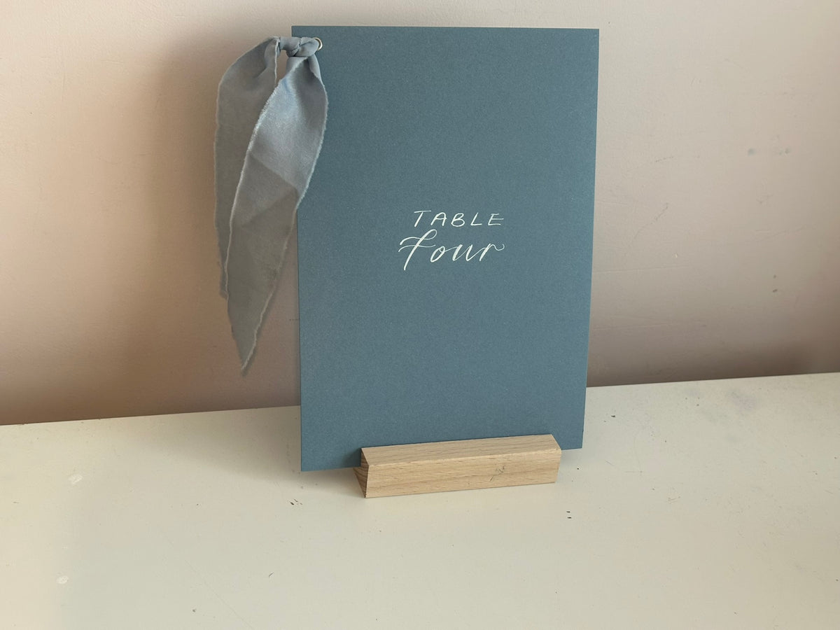 Mixed lettering wedding table number sign on dusky blue card with chiffon ribbon and silver calligraphy