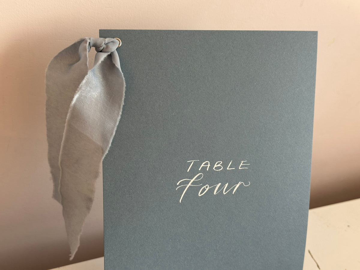 Mixed lettering wedding table number sign on dusky blue card with chiffon ribbon and silver calligraphy