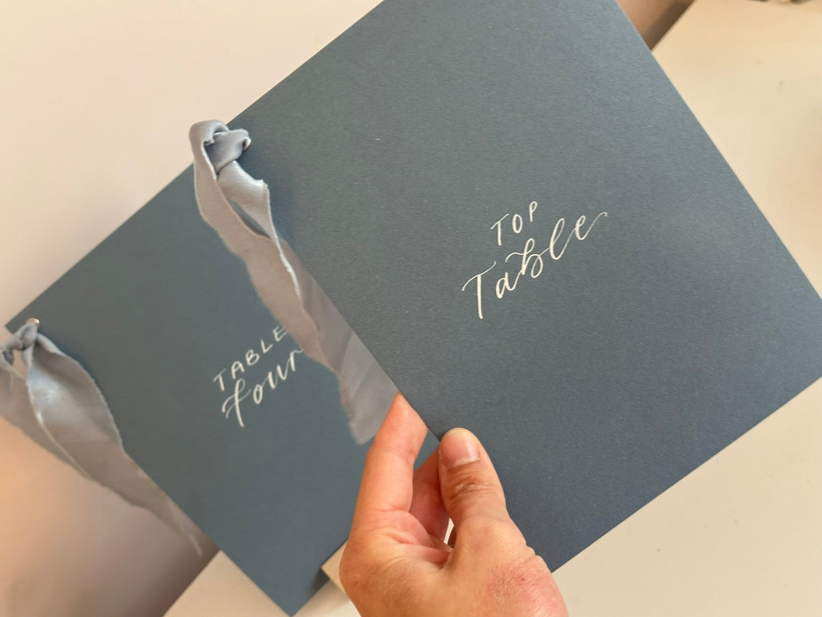 Mixed lettering wedding table number sign on dusky blue card with chiffon ribbon and silver calligraphy