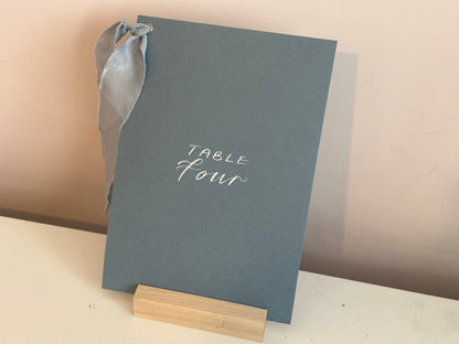 Mixed lettering wedding table number sign on dusky blue card with chiffon ribbon and silver calligraphy