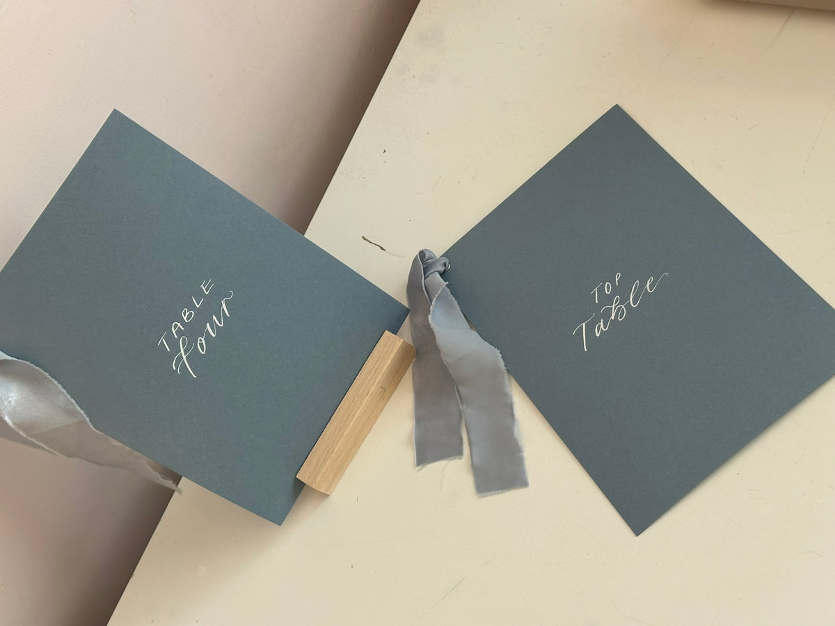 Mixed lettering wedding table number sign on dusky blue card with chiffon ribbon and silver calligraphy