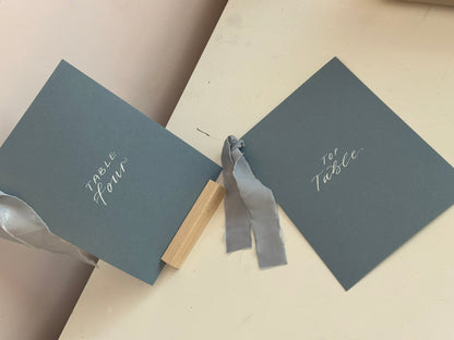 Mixed lettering wedding table number sign on dusky blue card with chiffon ribbon and silver calligraphy