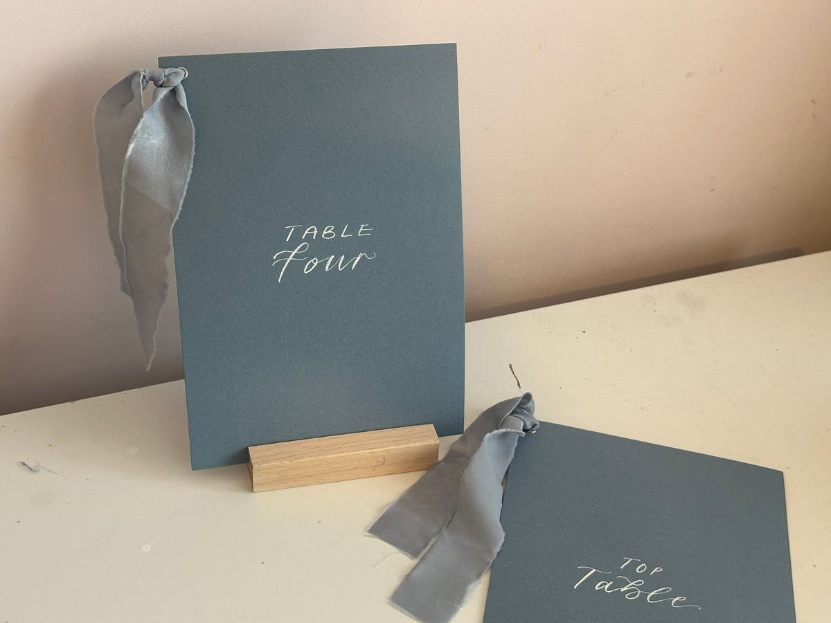 Mixed lettering wedding table number sign on dusky blue card with chiffon ribbon and silver calligraphy