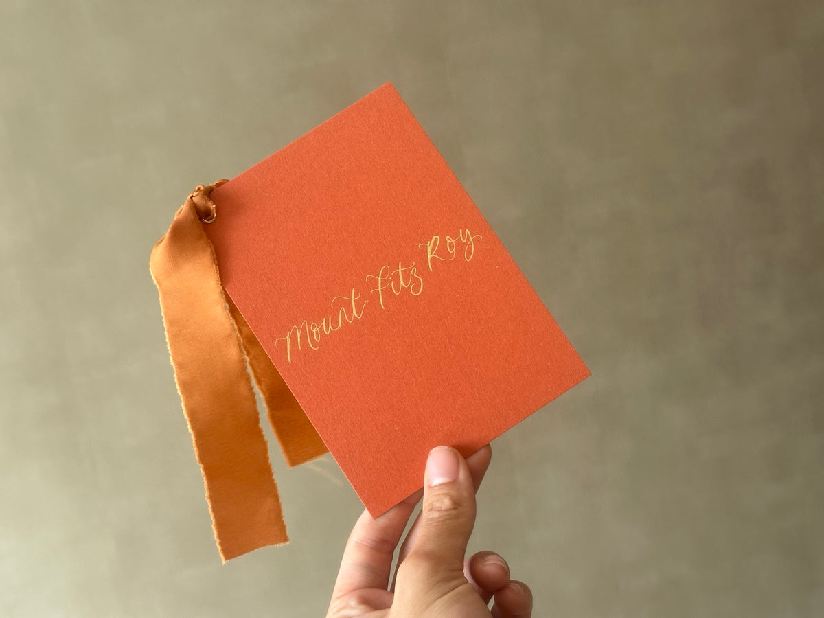 Rust orange wedding table name sign held in hand with gold calligraphy and ribbon