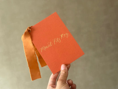 Rust orange wedding table name sign held in hand with gold calligraphy and ribbon
